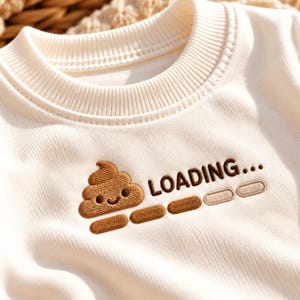 May include: A cream-colored long-sleeve shirt featuring an embroidered brown smiling poop emoji next to the text LOADING... and a horizontal progress bar consisting of four rectangular segments, with two segments filled in brown.