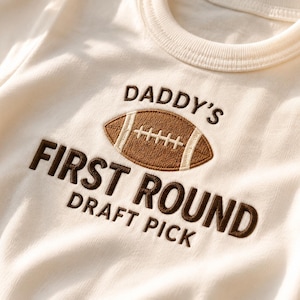 May include: An off-white short-sleeved shirt featuring brown embroidered text that reads DADDY'S FIRST ROUND DRAFT PICK. Centered above the text is an embroidered brown and white American football graphic with detailed stitching.
