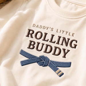 以下が含まれることがあります： DADDY'S LITTLE ROLLING BUDDYというダークブラウンの刺繍文字が入ったオフホワイトの半袖Tシャツ。文字の下には、結び目のある青い武道用ベルトが刺繍されており、ベルトの端には小さなベージュのストライプがあしらわれています。