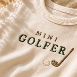 May include: A cream-colored long-sleeve sweatshirt featuring the embroidered text MINI GOLFER in dark green and brown thread. Below the text, there is an embroidered golf ball and a golf club in brown thread, centered on the chest area.