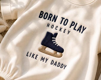 May include: A cream-colored short-sleeve bodysuit featuring dark blue embroidered text that reads BORN TO PLAY HOCKEY LIKE MY DADDY, with a central dark blue and tan embroidered ice skate graphic positioned between the lines of text.