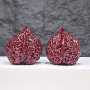 May include: Two dark red, glossy, walnut-shaped decorative objects with a highly textured, pitted surface. Each object features a prominent central ridge running vertically from the pointed top to the rounded base, mimicking the natural shell.