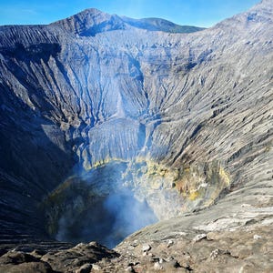 May include: A wide volcanic crater with steep, textured gray slopes and deep ridges leading down to a central vent. Wisps of white smoke rise from the bottom, where patches of yellow sulfur are visible against the dark, rocky terrain under a clear blue sky.