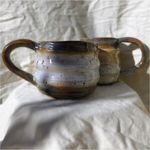 May include: Two handmade ceramic mugs with a rounded, ribbed shape and a glossy glaze. The front mug features a blend of lavender, white, and dark brown tones with speckles, while the back mug displays earthy tan and brown hues.