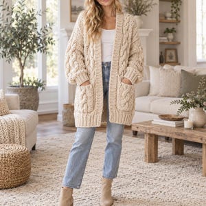 May include: A cream-colored, chunky cable-knit cardigan featuring an open front, long sleeves with ribbed cuffs, and two large front patch pockets. The garment is worn over a white tank top, light blue denim jeans, and tan ankle boots.