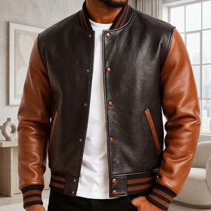 Op de afbeelding: Een varsity-stijl jack met een donkerbruin leren body en contrasterende lichtbruine leren mouwen. Het jack heeft een geribbelde kraag en boorden met bruine strepen, een sluiting met drukknopen aan de voorzijde en twee zijzakken.
