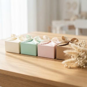 Könnte beinhalten: Vier quadratische Geschenkboxen in Creme, Mintgrün, Hellrosa und braunem Kraftpapier, jeweils mit einem gewellten Rand am Deckel und einer passenden Satinschleife. Ein kleines Bündel getrockneter weißer Blumen liegt neben der braunen Box.