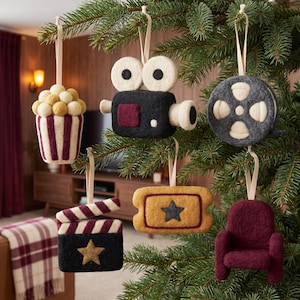 Op de afbeelding: Een set van zes vilten ornamenten hangend in een dennenboom, waaronder een popcornemmer, een zwarte filmcamera, een grijze filmrol, een zwart-bordeauxrode filmklapper met een gouden ster, een gouden ticket en een bordeauxrode bioscoopstoel.