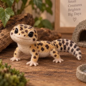 Könnte beinhalten: Eine nadelgefilzte Leopardgecko-Figur mit cremefarbenem Körper und dunkelbraunen Flecken. Der Gecko hat große schwarze Augen, ein breites lächelndes Maul und einen dicken Schwanz. Er steht auf einer strukturierten Oberfläche neben einer Steinhöhle.