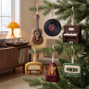Könnte beinhalten: Eine Sammlung von sechs Filzanhängern an einem Tannenbaum, darunter eine braune Akustikgitarre, eine schwarze Schallplatte, ein braunes Klavier, ein gelbes Radio, ein kastanienbraunes Metronom und eine grüne Kassette, jeweils mit cremefarbener Schlaufe.