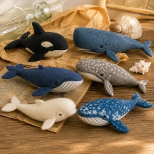 Puede incluir: Una colección de seis figuras de ballenas de lana fieltrada, incluyendo una orca, un cachalote, una beluga y tres ballenas barbadas con distintos patrones de color azul, gris y blanco, cada una con pequeños ojos de cuentas negras y lana.