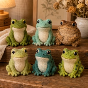 May include: Six needle-felted frog figurines arranged in two rows. The top row features a bright green frog, a pale mint green frog, and a brown speckled frog. The bottom row includes a light yellow-green frog, a teal frog, and a lime green frog.