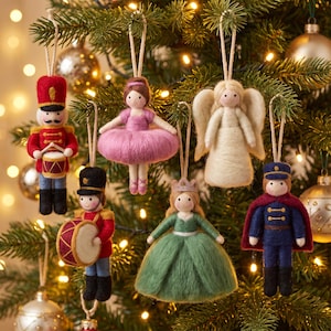 Könnte beinhalten: Ein Set aus sechs Weihnachtsanhängern aus Filz, die an einem Baum hängen. Die Figuren umfassen einen roten Soldaten mit Trommel, eine Ballerina im rosa Tutu, einen Engel mit weißen Flügeln, einen Trommler in Rot, eine Prinzessin im grünen Kleid und einen Prinzen.