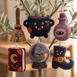 May include: A collection of six felted wool ornaments hanging from branches, including a broom, a cauldron, a potion bottle, a book with a crescent moon, a crystal ball, and a boot, each featuring a cartoon face with eyes and small teeth.
