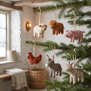 May include: A collection of six felt animal ornaments hanging from evergreen branches. The set includes a white sheep, a brown highland cow, a pink pig, a red chicken, a gray donkey, and a light gray goat, each suspended by a simple white cord.