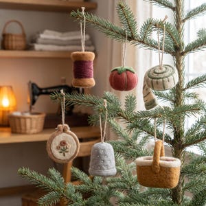 May include: A collection of six felt ornaments hanging from a pine tree, including a brown thread spool, a red tomato pincushion, a coiled measuring tape, an embroidery hoop with floral stitching, a grey thimble, and a tan woven basket.