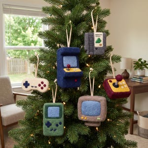 Peut inclure: Un ensemble de six décorations en feutrine représentant des consoles de jeux rétro, une console portable, un écran d'ordinateur, une cartouche de jeu et une manette, suspendues par des ficelles blanches sur un sapin de Noël artificiel.