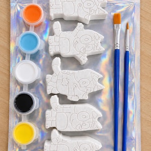 May include: A craft kit featuring five white plaster figures of a cartoon dog, a strip of five small paint pots in orange, blue, white, black, and yellow, and two blue-handled paintbrushes, all packaged inside a holographic silver plastic pouch.