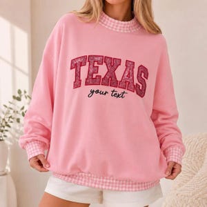 May include: A light pink long-sleeve sweatshirt featuring a mock neck collar and cuffs with a white and pink gingham pattern. The front displays the word TEXAS in large, glittery red letters with the phrase your text written below in black.