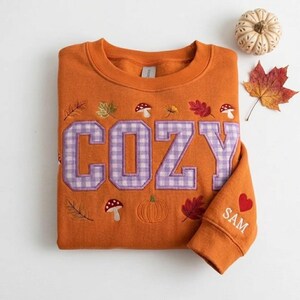 Puede incluir: Una sudadera naranja de cuello redondo con la palabra COZY en letras grandes de cuadros vichy de color lila claro. El diseño incluye setas, calabazas y hojas de otoño bordadas. La manga tiene un corazón rojo y el nombre SAM bordado en blanco.