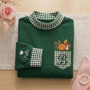 May include: A dark green long-sleeve shirt with a mock neck collar featuring a green and white gingham pattern. The shirt has a matching gingham pocket with an embroidered pumpkin and mushroom design, a large letter B, and a sleeve with the name Bella.