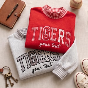 May include: Two long-sleeve sweatshirts, one red and one light gray, featuring the word TIGERS in large block letters with a mix of gingham and polka dot patterns. Below the word TIGERS, the text your text is embroidered in a cursive font.