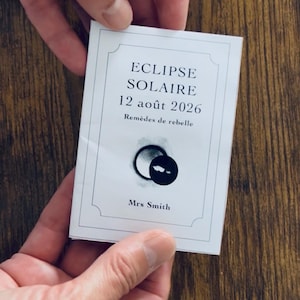 Peut inclure: Une carte rectangulaire blanche avec une fine bordure noire portant le texte ECLIPSE SOLAIRE, 12 août 2026, Remèdes de rebelle et Mrs Smith. Sous le texte se trouve une illustration graphique d'une éclipse solaire noire sur fond blanc.