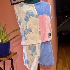 Peut inclure: Un ensemble deux pièces composé d'un haut sans manches et d'un short en tissu éponge. Le vêtement présente un design color-block avec des sections à motif floral sur fond crème, ainsi que des panneaux unis bleu clair et rose.