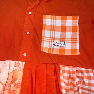 Peut inclure: Une chemise boutonnée orange uni avec une poche poitrine à carreaux orange et blancs portant une petite étiquette indiquant cherry. La partie inférieure de la chemise est composée de panneaux en tissu orange, à carreaux et à motifs floraux.