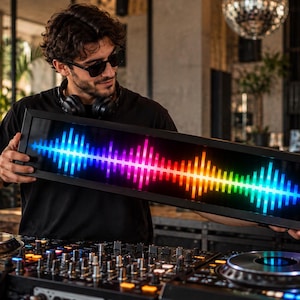 May include: A rectangular black-framed LED display panel featuring a vibrant, multi-colored digital audio waveform graphic that transitions through shades of blue, purple, pink, orange, and yellow across the horizontal screen surface.