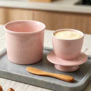 Peut inclure: Un beurrier en céramique rose clair avec le mot BUTTER en relief sur le côté, un couvercle rose assorti contenant un bloc de beurre jaune pâle et un couteau à beurre en bois lisse posés sur un plateau rectangulaire en pierre grise.