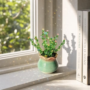 May include: A decorative cactus sculpture crafted from faceted green glass beads with small pink accents, potted in a rounded, light green ceramic vessel with a subtle crackle glaze finish, positioned near two books titled Wabi-Sabi Welcome.
