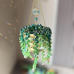 Op de afbeelding: Een hangend plantornament met een klein gevlochten mandje gevuld met trapsgewijze strengen van gefacetteerde groene kristallen kralen. Het object hangt aan gouden kettingen en bevat een ronde bedel met geperste blauwe bloemen en een groene kraal.
