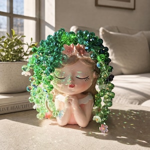 May include: A ceramic planter shaped like a person with closed eyes and hands clasped together. The figure is adorned with a cascading arrangement of faceted green crystal beads that mimic trailing vines, accented with small pink and white flowers.