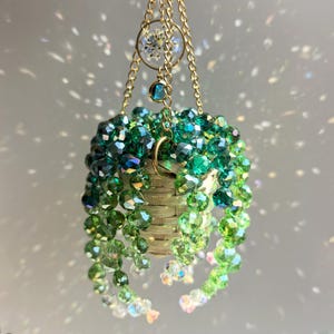 May include: A hanging ornament featuring a miniature woven basket filled with cascading strands of faceted green crystal beads. The basket is suspended by gold-toned chains, with a small circular charm containing a dried white flower.