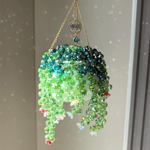 Op de afbeelding: Een hangend plantornament gemaakt van gefacetteerde groene en blauwgroene kristallen kralen, gerangschikt in trapsgewijze strengen. De plant zit in een kleine witte pot die aan goudkleurige kettingen hangt, met een kleine ronde bedel.