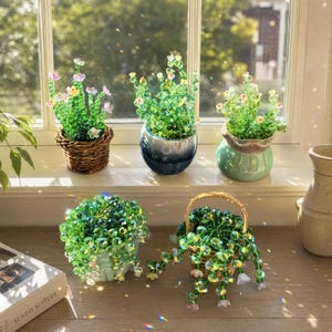 May include: Five decorative potted plants crafted from green and clear glass beads. Three upright plants sit in a woven basket, a blue ceramic pot, and a light green pot. Two trailing plants are in a light green pot and a small woven basket.