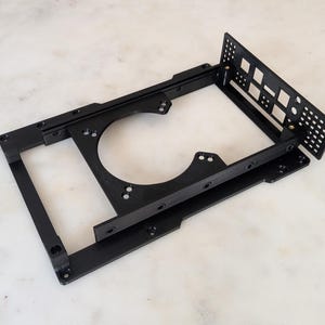 Può includere: Una staffa di montaggio in metallo nero per componenti informatici, caratterizzata da un telaio rettangolare con un'apertura circolare centrale e un pannello laterale perforato perpendicolare con fori quadrati.