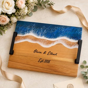May include: A rectangular wooden serving tray featuring a resin ocean wave design with deep blue and white foam accents. The tray includes black metal handles on each side and is engraved with the text Olivia & Ethan and Est. 2026.
