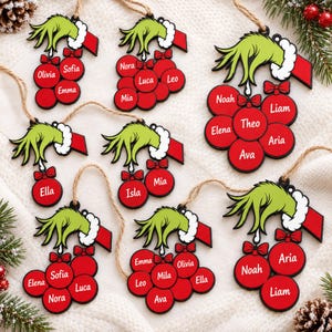 Op de afbeelding: Een collectie kerstornamenten met een groene harige hand met een witte manchet die trossen rode ronde kerstballen vasthoudt. Elke kerstbal is gepersonaliseerd met een witte naam en hangt aan een bruin touwtje.