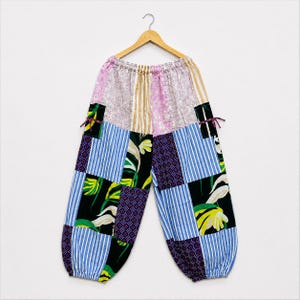 Può includere: Pantaloni patchwork con elastico in vita e un mix di tessuti tra cui righe blu e bianche, motivi floreali neri con fiori gialli e bianchi e stampe geometriche viola. I pantaloni presentano tasche laterali con piccoli laccetti di chiusura.