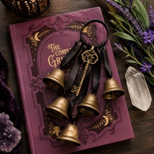 Può includere: Un ornamento decorativo da appendere composto da un anello in metallo nero decorato con cinque campanelli color ottone e una chiave antica centrale, il tutto sospeso da nastri di raso nero, appoggiato su un libro viola intitolato The Complete Grimm.