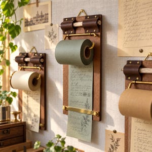 May include: A wall-mounted paper roll dispenser featuring a dark brown wooden board, a brass metal holder, and dark brown leather straps. The dispenser holds a roll of sage green paper that extends downward, displaying printed cursive text and botanical illustrations.