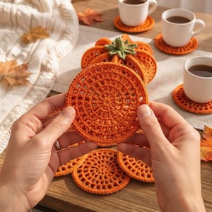 Puede incluir: Un posavasos redondo de ganchillo naranja con un patrón de puntos circulares texturizados. El posavasos se sostiene en primer plano, mientras que otros posavasos de ganchillo naranjas a juego y una calabaza de ganchillo se ven al fondo.