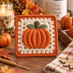 Puede incluir: Un cuadrado de ganchillo que presenta una calabaza naranja central con un tallo marrón y dos hojas verdes. La calabaza está rodeada por un borde texturizado de color crema y rematada con un borde exterior de puntos de hilo naranja.