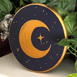 May include: A circular wooden plaque painted with a dark navy blue background featuring a large, textured gold crescent moon at the centre. The design includes small gold painted dots representing stars and a four-pointed star, with a gold painted rim.