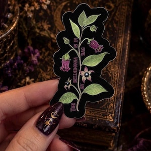 May include: A die-cut sticker featuring a botanical illustration of a belladonna plant with green leaves and purple bell-shaped flowers. The text "THE BELLADONNA IS IN MY BAG" is printed vertically along the stem in a dark purple font on a black background.