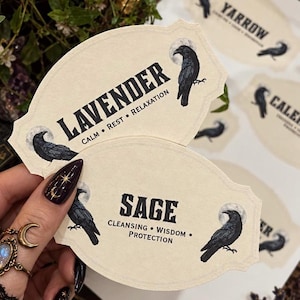 May include: Two cream-coloured paper labels with black text and raven illustrations. One label reads LAVENDER CALM REST RELAXATION, the other reads SAGE CLEANSING WISDOM PROTECTION. Each label features a black raven perched near a full moon graphic.