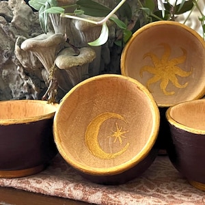 May include: A set of small, round wooden bowls with dark brown exterior walls and natural light wood interiors. Each bowl features a gold painted rim and a gold celestial symbol at the base, including a crescent moon with a star and a sun design.