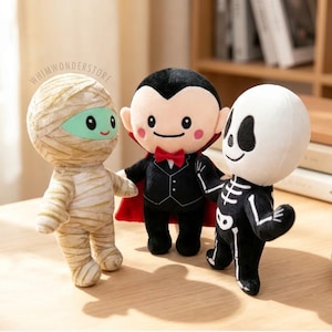 Puede incluir: Tres peluches que incluyen una momia envuelta en vendas beige con cara verde, un vampiro con traje negro, capa roja y pajarita, y un esqueleto con cráneo blanco y cuerpo negro. El texto WHIMWONDERSTORE es visible en la parte superior.